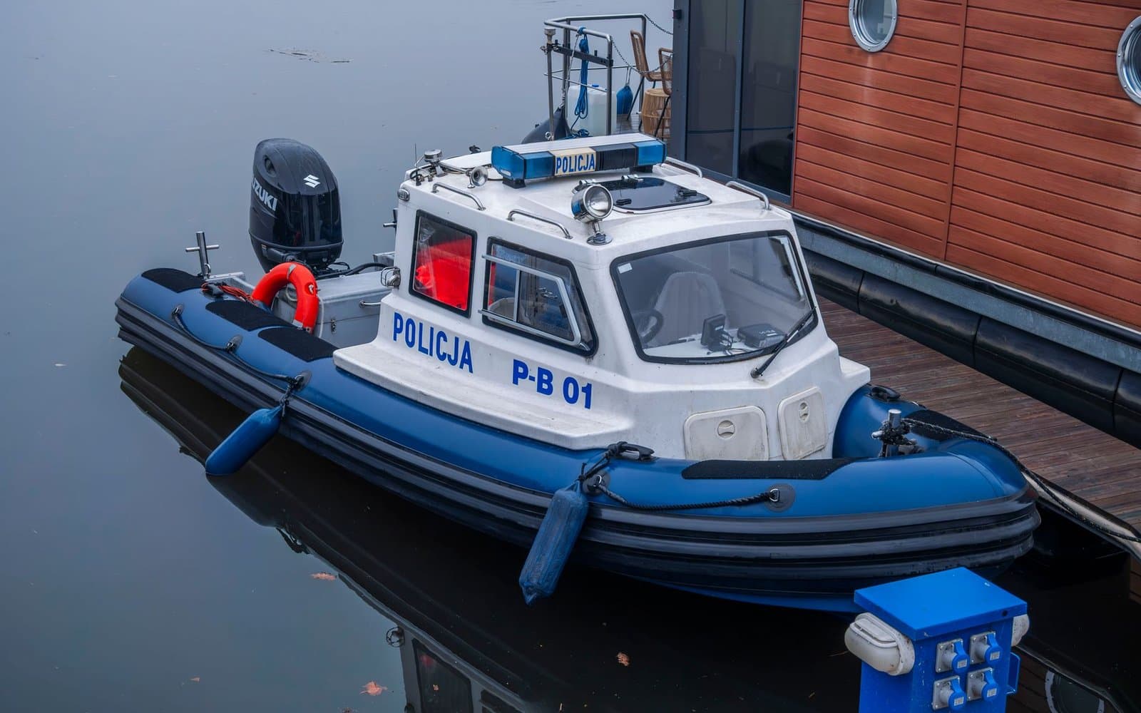 Policyjna łódź patrolowa z napisem POLICJA zacumowana przy pomoście przystani, obok drewniany pomost i bosman pływający